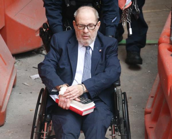 Harvey Weinstein in a suit and tie, sitting in a wheelchair, being escorted by armed police officers out of Bellevue Hospital in New York
